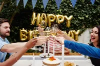 Friends gathered around a table raising glasses of white wine in a toast at an outdoor birthday party, with a cake and candles on the table and golden 'Happy Birthday' balloons in the background.