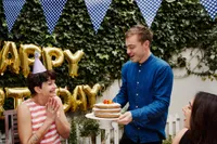 Smiling guest wearing a party hat claps with excitement as another person presents a layered birthday cake with candles at an outdoor celebration, with 'Happy Birthday' balloons and bunting behind them.