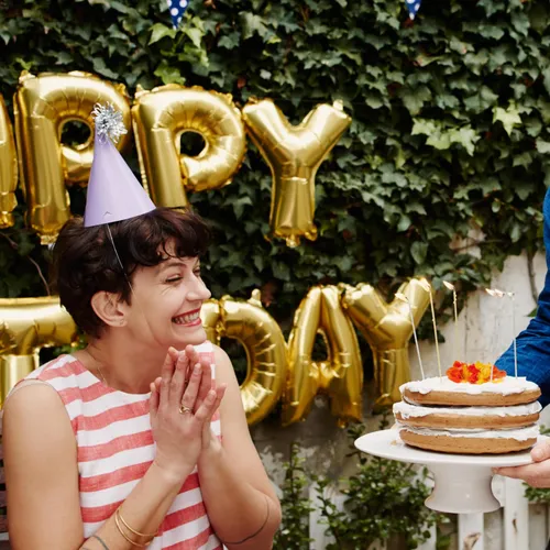 Person in a striped shirt wearing a party hat smiles as a birthday cake with lit candles is presented; gold 'Happy Birthday' balloons hang behind.