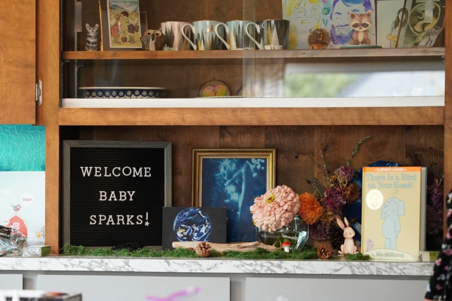 A letter board reading "Welcome Baby Sparks!" beside zinnias, a framed cyanotype, a ceramic rabbit, and a children's book.