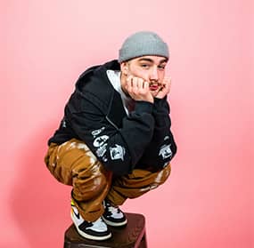 Young man crouching on a box, wearing a gray beanie, black hoodie, and patterned brown pants, resting his face on his hands and looking toward the logo in the center.