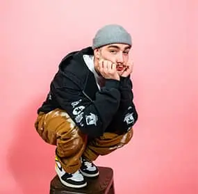 Young man crouching on a box, wearing a gray beanie, black hoodie, and patterned brown pants, resting his face on his hands and looking toward the logo in the center.