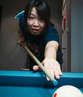 Woman aiming a pool cue toward the camera, focused on her shot, while glancing toward the logo in the center with a determined expression.