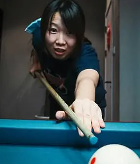 Woman aiming a pool cue toward the camera, focused on her shot, while glancing toward the logo in the center with a determined expression.