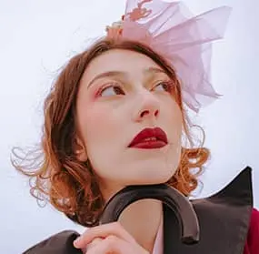 Woman with short red hair, wearing a pink headpiece and a black outfit, looking thoughtfully upward and toward the logo in the center.
