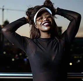 Smiling woman with curly hair in a visor and dark shirt, standing outdoors, arms raised behind her head, joyfully looking toward the logo in the center.