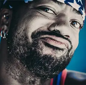 Close-up of a man with tattoos, a nose ring, and a bandana giving a playful side-eye, looking toward the logo in the center with a mischievous expression.