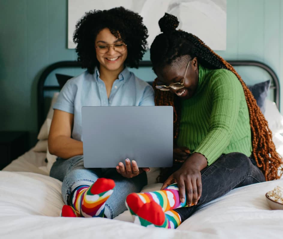 Two women sitting on a bed wearing rainbow socks, smiling while looking at a laptop together.